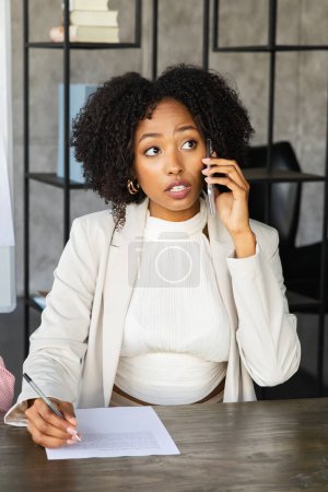 Foto de Emprendimiento femenino. Retrato vertical de la exitosa joven mujer de negocios negra con traje formal sentada en el escritorio, firmando papeles, hablando por teléfono con su socio o cliente - Imagen libre de derechos