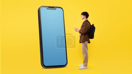 Photo for Student Guy Using His Smartphone Looking At Huge Cellphone With Empty Screen, Websurfing Information About New Innovative Application Standing With Backpack In Studio On Yellow Background - Royalty Free Image