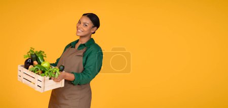 Photo for Garden Harvest. Happy Black Farmer Lady Holding Wooden Box With Fresh Ripe Vegetables, Fruits And Herbs Standing Over Yellow Studio Background, Smiling To Camera. Panorama With Empty Space - Royalty Free Image