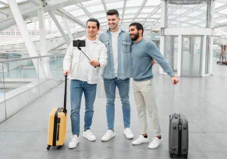 Foto de Equipo de Viaje Aéreo. Tres bloggers hombres con maletas haciendo selfie en el teléfono inteligente en la puerta de salida en el aeropuerto moderno. Grupo de Pasajeros Divirtiéndose Esperando Su Vuelo. Longitud completa - Imagen libre de derechos