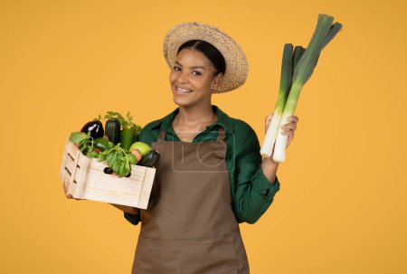 Photo for Successful Gardening. Smiling Black Lady Farmer Holding Leek And Wooden Box With Fresh Farm Vegetables Posing Wearing Summer Hat Over Yellow Studio Background. Harvest Time Concept - Royalty Free Image