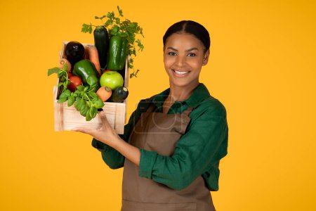 Foto de Oferta de comida ecológica. Mujer agricultora negra mostrando su cosecha de jardín sosteniendo una caja de madera con verduras y frutas frescas, sonriendo a la cámara posando sobre fondo amarillo, fotografía de estudio - Imagen libre de derechos