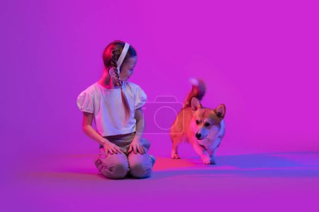Téléchargez les photos : Portrait de belle petite fille préadolescente portant une tenue d'été élégante et un casque sans fil jouant avec un chien chiot gallois mignon, fond coloré avec effet lumineux, espace de copie - en image libre de droit