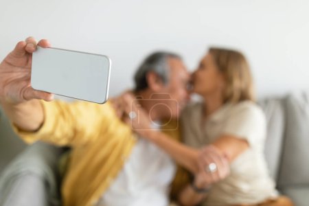 Téléchargez les photos : Aimer femme caucasienne âgée embrasser mari et embrasser, homme faisant selfie sur smartphone, assis sur le canapé, foyer sélectif. Photo pour les réseaux sociaux et blog, amour et relation à la maison - en image libre de droit