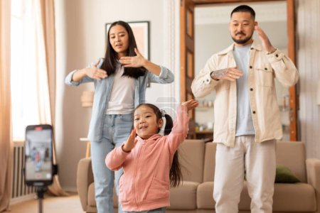Téléchargez les photos : Bloguer et les médias sociaux. Famille japonaise joyeuse avec petite fille filmant leur danse par téléphone, faire de la vidéo et s'amuser ensemble à la maison. Focus sélectif sur l'enfant - en image libre de droit
