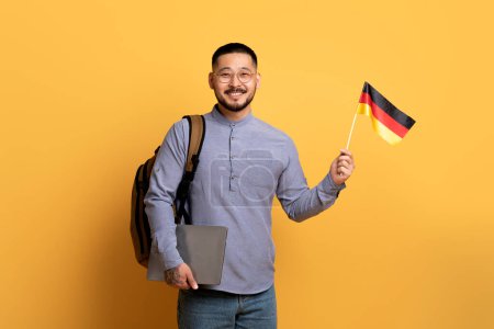 Foto de Cursos de idiomas en línea. Sonriente joven asiático hombre sosteniendo bandera alemana y el ordenador portátil, mientras que de pie sobre el fondo amarillo en el estudio, Millennial hombre recomendando programas de educación remota, Copiar el espacio - Imagen libre de derechos