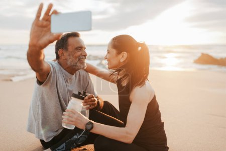 Photo for Glad mature caucasian couple in sportswear rest from fitness, sit on sand with bottles of water, take selfie on smartphone at sea beach, outdoor. Workout blog, health care, app for body care - Royalty Free Image