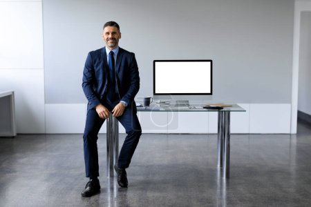 Photo for Handsome middle aged businessman in formal suit sitting near office table with blank computer monitor, offering mockup for your design, free space. Business ad concept - Royalty Free Image