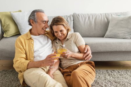 Téléchargez les photos : Aimer les conjoints âgés boire du jus d'orange frais, embrasser et profiter du temps ensemble à la maison, espace libre. Couple marié âgé assis profiter d'une boisson saine, câlins et célébrer l'anniversaire - en image libre de droit