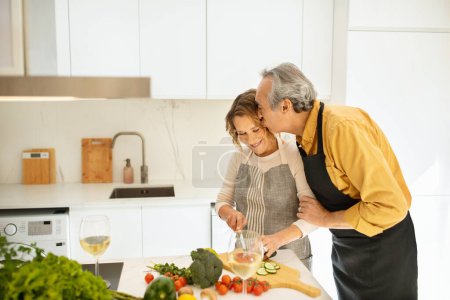 Foto de Amar marido mayor abrazar y besar a su cónyuge sonriente en la acogedora cocina, mujer cocinar cena saludable para su familia en casa, espacio de copia - Imagen libre de derechos