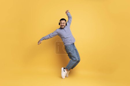 Téléchargez les photos : Joyeux homme asiatique portant des écouteurs sans fil dansant sur fond jaune, beau jeune homme millénaire dans les lunettes de vue s'amuser, écouter de la musique sur Smartphone et debout sur ses orteils - en image libre de droit