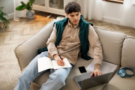 Photo for E-Learning Offer. Arabic guy having online class on laptop in living room, taking notes during lecture at home. Student doing homework engaging in distance education leisure - Royalty Free Image