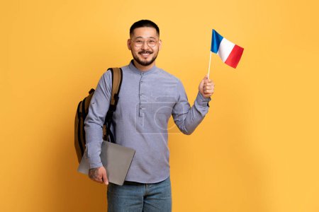 Photo for Study Abroad. Asian Man With Backpack Holding Laptop And Flag Of France, Smiling Millennial Male Student Enjoying Learning French Language, Standing Over Yellow Background In Studio, Copy Space - Royalty Free Image
