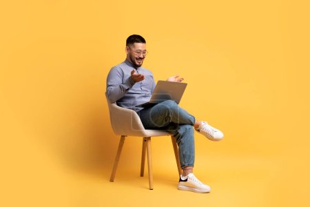 Photo for Cherful Young Asian Man Making Video Call On Laptop While Sitting In Armchair Over Yellow Studio Background, Excited Millennial Guy Looking At Computer Screen And Talking At Web Camera, Copy Space - Royalty Free Image