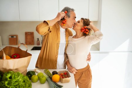Téléchargez les photos : Aimer les conjoints aînés arrivant du supermarché avec sac d'épicerie et déballer, s'amuser avec des tomates cerises et s'embrasser à l'intérieur de la cuisine - en image libre de droit