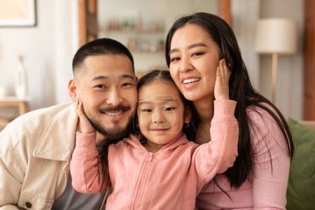 Téléchargez les photos : Unité familiale. Portrait d'une belle famille coréenne de trois embrassant exprimant des émotions positives posant à la maison. Les parents étreignant leur adorable fille enfant souriant à la caméra ensemble - en image libre de droit