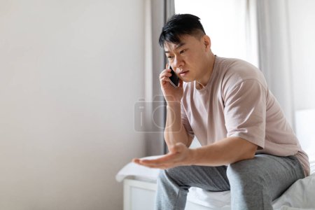 Photo for Angry emotional handsome middle aged asian man sitting on bed at home, have phone conversation, fighting with lover or business partner, wearing pajamas, copy space. Communication concept - Royalty Free Image