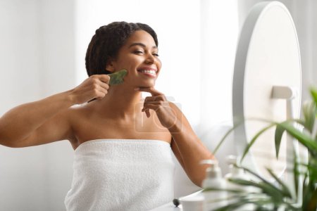 Téléchargez les photos : Beauty Tools. Jeune femme noire heureuse faisant massage du visage avec grattoir Gua Sha, femme afro-américaine souriante enveloppée dans une serviette après le bain faisant des traitements du visage à la maison, Copier l'espace - en image libre de droit