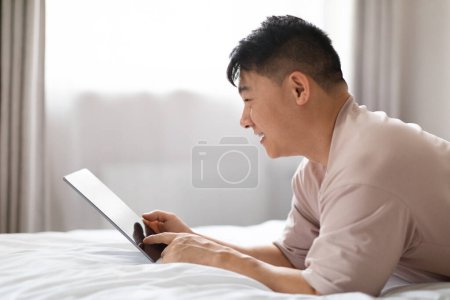 Téléchargez les photos : Application énergisante. Joyeux sourire bel homme asiatique d'âge moyen portant des vêtements de maison couché sur le lit à la maison, en utilisant une belle application sur tablette numérique, ebook de lecture, blog, défilement, espace de copie - en image libre de droit