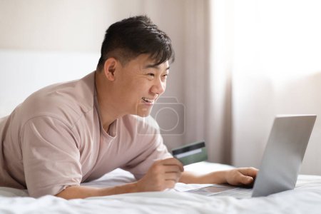 Téléchargez les photos : Relaxé heureux beau moyen âge asiatique homme portant pyjama bancaire sur Internet à partir du lit tout en passant week-end à la maison, en utilisant moderne PC portable et carte bancaire, copier l'espace - en image libre de droit