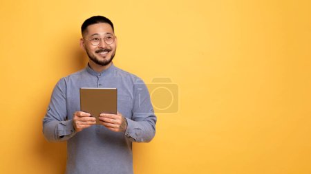 Foto de Joven sonriente hombre asiático sosteniendo la tableta digital y mirando hacia otro lado, pensativo soñador millennial masculino con gadget moderno en las manos de pie sobre fondo de estudio amarillo, pensando en la oferta agradable, Panorama - Imagen libre de derechos