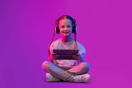 Photo for Digital Native, Generation Alpha Concept. Preteen kid happy pretty adorable girl in summer outfit sitting on floor over futuristic background, using wireless headphones and digital tablet, copy space - Royalty Free Image