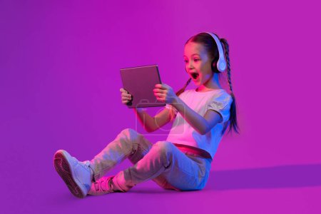 Téléchargez les photos : Apprentissage virtuel, nouvelle forme d'éducation interactive et numérique. Petite fille étonnante assise sur le sol, utilisant une tablette numérique et des écouteurs sur fond lumineux, profitant d'une application éducative, espace de copie - en image libre de droit