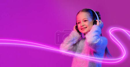 Téléchargez les photos : Digital Wonderland. Jolie petite fille utilisant des écouteurs sans fil, écouter des sons fantastiques du monde numérique, fond coloré avec lumière au néon. Magie et mystère de l'univers numérique, espace de copie - en image libre de droit