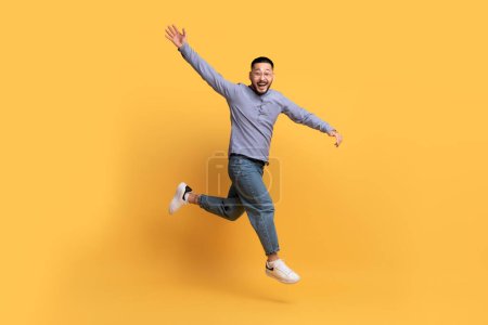 Téléchargez les photos : Heureux homme asiatique excité sautant dans les airs sur fond jaune, en mouvement prise de vue émotionnelle gai Millennial Guy en vêtements décontractés s'amusant en studio, pleine longueur prise de vue avec l'espace de copie - en image libre de droit