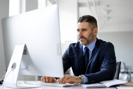 Téléchargez les photos : Homme d'affaires d'âge moyen en costume assis à la table de travail au bureau devant l'ordinateur, tapant sur le clavier, envoyant des e-mails à ses partenaires d'affaires, travaillant sur la recherche marketing, l'espace de copie - en image libre de droit