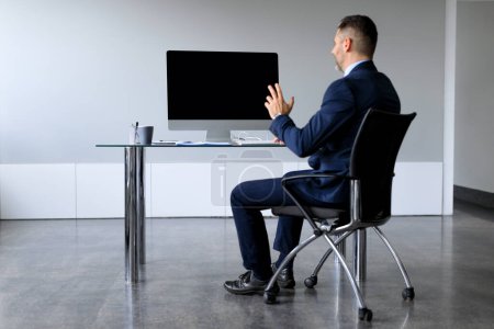 Photo for Rear view of middle aged businessman looking at blank empty computer monitor, having online web video call, sitting at desk at workplace, free copy space banner - Royalty Free Image