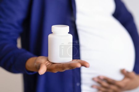 Foto de Joven mujer embarazada negro con el vientre grande muestra frasco de vitaminas, pastillas sobre fondo blanco de la pared, recortado. Atención de la salud, expectativa del niño en el hogar, tratamiento y recomendación - Imagen libre de derechos
