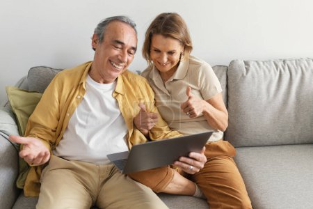 Téléchargez les photos : Couple caucasien âgé souriant ayant appel vidéo via ordinateur portable avec la famille, femme montrant pouces jusqu'à webcam, assis sur le canapé dans le salon. Distance sociale avec gadget - en image libre de droit