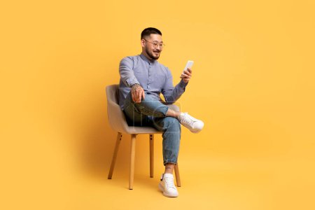 Photo for Great App. Young Asian Man Using Smartphone While Sitting In Chair Over Yellow Background, Happy Millennial Male Browsing New Mobile Application Or Messaging Online, Scrolling Social Networks - Royalty Free Image