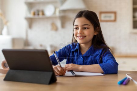 Photo for E-Learning. Happy Middle Eastern Schoolgirl Looking At Digital Tablet And Taking Notes, Excelling in Her Studies Through Online Education At Home, Doing Homewotk Sitting At Desk Near Computer - Royalty Free Image