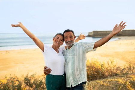Téléchargez les photos : Destination de vacances de rêve. Joyeux couple d'âge mûr levant les mains embrassant posant près de la plage de l'océan en plein air au Portugal, souriant à la caméra. Conjoints profiter de l'été à bord de la mer Gesturing Bonjour - en image libre de droit