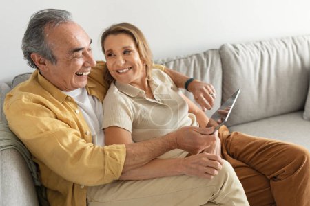 Téléchargez les photos : Heureux couple marié de personnes âgées profitant du monde gadget numérique à l'aide d'un ordinateur tablette, websurf ou regarder un film en ligne, reposant sur un canapé. Retraite et concept amusant d'Internet - en image libre de droit