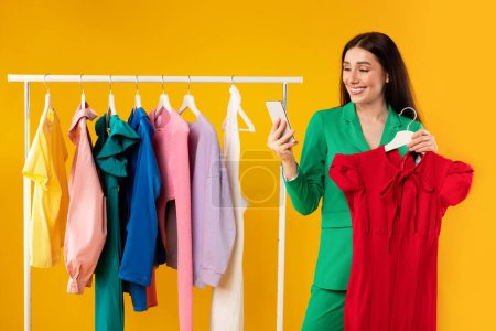 Photo for Happy smiling lady using smartphone and holding red dress, woman ordering clothes online in mobile application, posing over yellow studio background. App for online shopping - Royalty Free Image