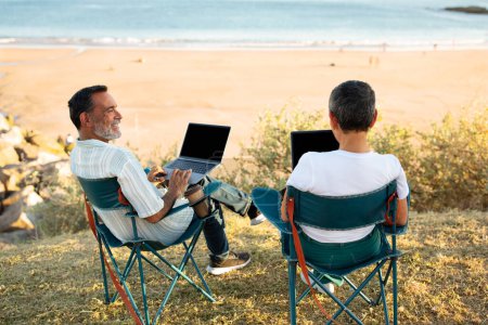 Téléchargez les photos : Freelance Dream. Vue arrière du couple aîné utilisant des ordinateurs portables assis dans des chaises à la plage près de l'océan en plein air. Mature conjoints naviguant sur Internet en vacances, appréciant le travail en ligne de n'importe où - en image libre de droit