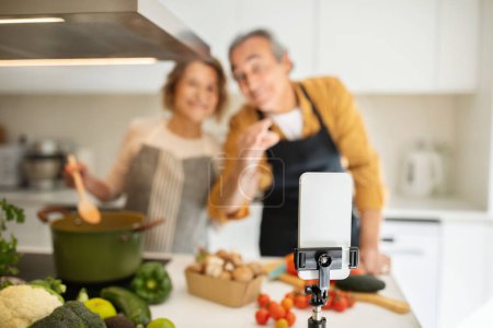 Photo for Selective focus on smartphone set on tripod over loving senior spouses cooking dinner together at modern kitchen, blurred background. Retirement and cooking blogging concept - Royalty Free Image