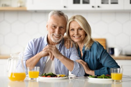 Photo for Portrait of happy mature couple posing in kitchen while eating lunch together, cheerful senior husband and wife sitting at table, embracing and smiling at camera, having delicious meal, free space - Royalty Free Image
