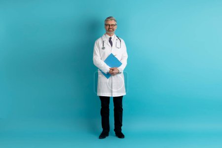Photo for Full length studio shot of cheerful friendly cheerful middle aged european doctor wearing eyeglasses holding medical chart posing on blue studio background, copy space. Medical staff, doctors - Royalty Free Image