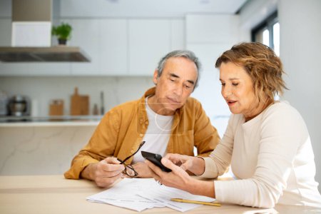 Photo for Senior couple sitting in kitchen with papers and bills and using calculator, husband and wife counting monthly expenses or planning family budget at home - Royalty Free Image