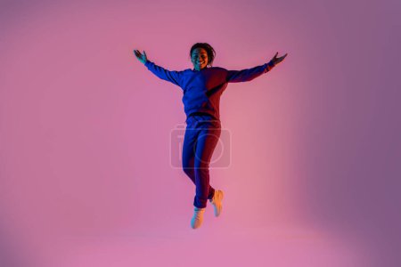 Foto de Mujer negra emocionada bailando y saltando sobre fondo rosa en luz de neón, mirando y sonriendo a la cámara, de cuerpo entero. Concepto de danza, juventud, afición, dinámica, movimiento, acción, anuncio - Imagen libre de derechos