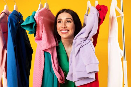 Photo for Smiling beautiful european woman posing between colorful new clothes hanging on garment rack, lady looking aside and smiling on yellow background. Shopping time concept - Royalty Free Image