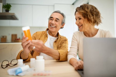 Los cónyuges caucásicos envejecidos que compran medicamentos en línea, pareja de personas mayores que buscan receta para pastillas con la ayuda de la computadora portátil, sentado en la mesa en el interior de la cocina