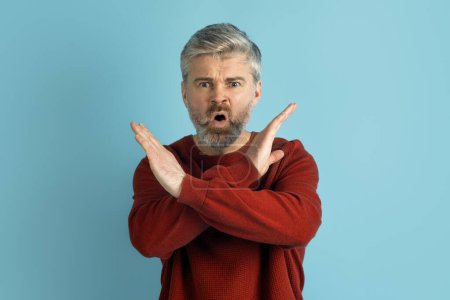 Photo for Image of angry middle aged man looking serious, making cross hands gesture to stop something bad, telling no, prohibit action, standing over blue studio background. Gestures and emotions concept - Royalty Free Image