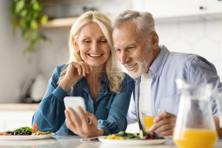 Téléchargez les photos : Heureux couple de personnes âgées utilisant un smartphone tout en prenant le petit déjeuner à la cuisine, gai homme et femme âgés naviguant sur Internet sur le téléphone mobile ou vérifier les photos sur les médias sociaux tout en mangeant de la nourriture - en image libre de droit