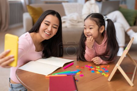 Photo for First Lesson. Cheerful Japanese Mom Making Selfie On Phone Teaching Little Preschool Daughter To Read And Write Sitting At Table At Home. E-Learning And Early Education Activities. Selective Focus - Royalty Free Image