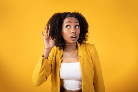 Photo for Overhearing, gossips and interesting news. Curious black curly lady listening secrets eavesdropping holding hand near ear posing over yellow studio background - Royalty Free Image